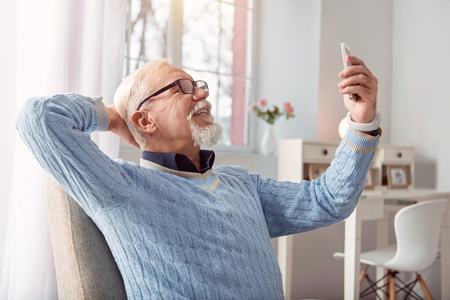 Making good memory. Pleasant upbeat senior man sitting in the armchair and posing for the camera, smiling widely, while taking selfiesの写真素材