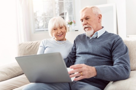Keeping themselves updated. Pleasant elderly husband and wife sitting on the couch and scrolling the...