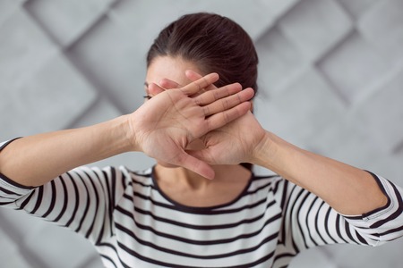 Do not look. Nice pleasant young woman standing and holding her hands together while covering her faceの写真素材