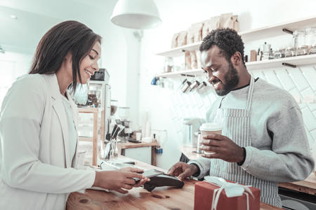 Your order. Nice positive delighted man standing at the counter and holding a cup of coffee while giving it to the customerの写真素材
