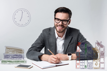 Working day. Positive experienced smiling man feeling glad while sitting in a comfortable office and making notes in his personal notebookの写真素材