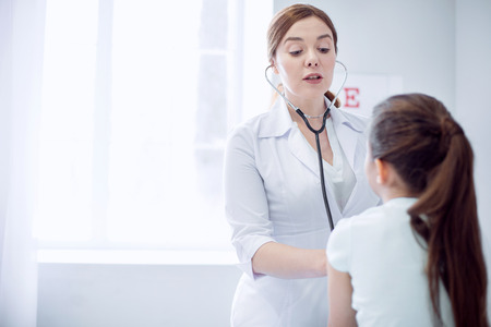 Let me hear. Beautiful appealing female doctor hearing girl while using stethoscope and standing on light backgroundの写真素材