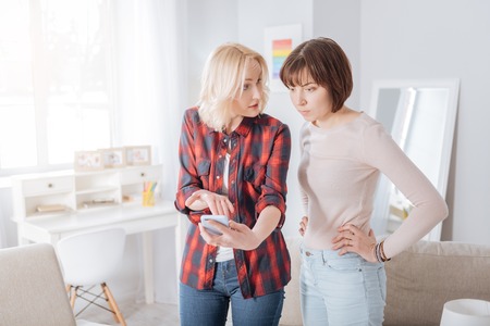 Look at this. Nice upset cheerless woman holding a smartphone and pointing at it while having a quarrel with her girlfriendの写真素材
