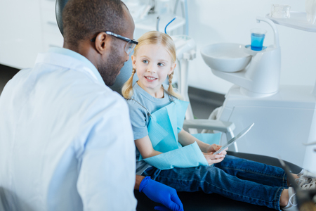 Pleasant pastime. Adorable little girl using tablet and talking to her dentist while having an appointment with himの写真素材
