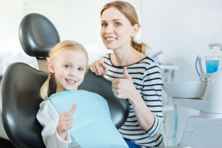 Satisfied with service. Pretty little girl and her beloved mother showing thumbs up while the girl sitting in the dentist chair and waiting for the treatmentの写真素材