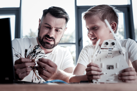 Two peas in a pod. Selective focus on a concentrated parent looking at wires while sitting next to his cheerful son and working on a new self automated robot together.の写真素材