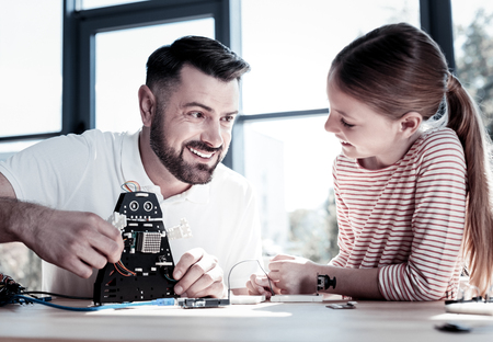 So proud of her. Selective focus on a joyful parent smiling cheerfully while looking at his smart curious daughter and constructing a new robot together.の写真素材