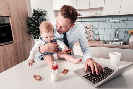 Keep calm. Serious man holding his kid and keeping left hand on the keyboard while trying to workの写真素材