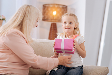 Joyful nice cute girl smiling and holding a gift box while being together with her grandmotherの写真素材