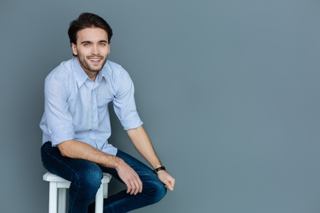 Positive emotions. Delighted cheerful bearded man sitting on a stool and smiling to you while being in a great moodの写真素材