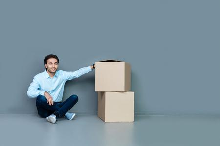 Cardboard boxes. Nice positive delighted man sitting on the floor and smiling while putting his hand on cardboard boxesの写真素材