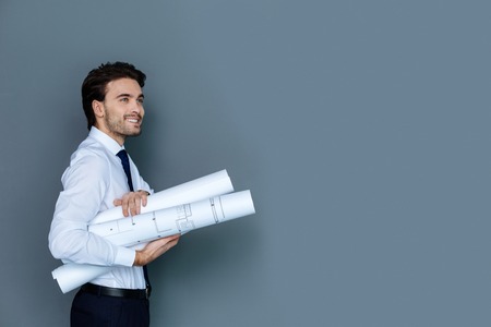 Professional drawings. Joyful positive smart man smiling and holding blueprints while working in the officeの写真素材