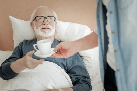 Senior unshaken bespectacled man lying on the bed looking up and taking a cup.の写真素材
