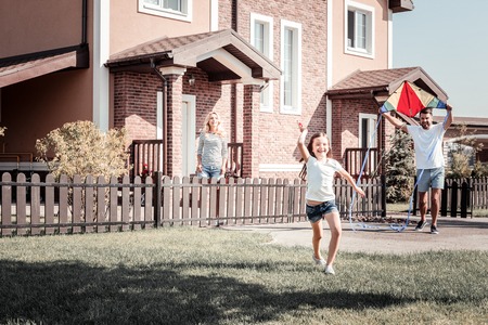 Daddy play with me. Joyful little smiling girl playing with near their home on the yard holding the air kite dad and running.の写真素材