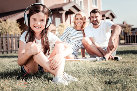 Favorite song. Little pretty satisfied girl sitting on the ground near her parents smiling and listening to music.の写真素材
