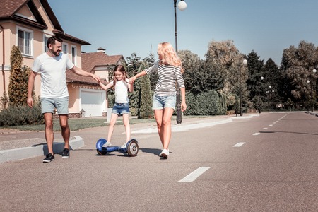 You will be next. Reliable satisfied lovely parents supporting theirs daughter who riding on the self-balancing scooter holding her by hands and smiling.の写真素材