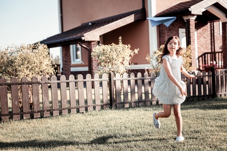 Fly far. Concentrated cute pleasant girl playing on the yard running and throwing a paper plane.の写真素材