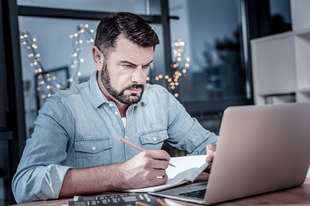 Its important thing. Busy responsible qualified sitting on his workplace by the desktop worker looking at his laptop and writing.の写真素材