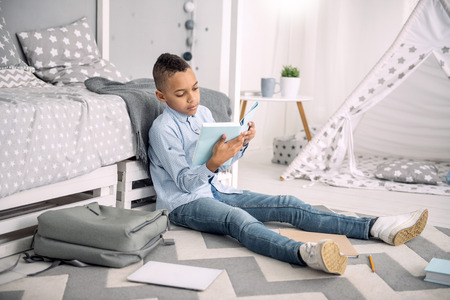School program. Appealing afro american boy sitting on floor while studying bookの写真素材
