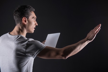 Progressive technology. Intelligent nice brunette man holding a tablet and stretching out his hand while working with a computer sensory panelの写真素材