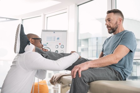 Medical assistance. Professional smart doctor holding his patients leg while providing medical...