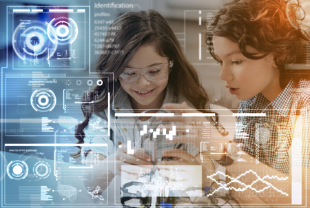 Exciting activity. Little clever girl wearing special glasses and smiling while her serious friend helping with making a robot and looking surprisedの写真素材