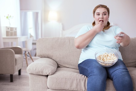 Bad habits. Dreamy red-head woman sitting on the sofa eating popcorn while watching televisionの写真素材