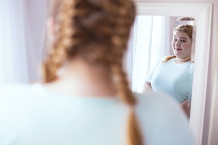 Body positivity. Smiling plump woman wearing turquoise sport shirt standing near mirrorの写真素材