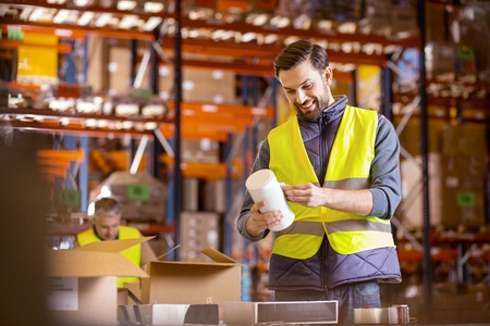 Factory production. Joyful warehouse worker wearing special uniform while putting sticker on...