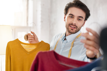 Great look. Cheerful bearded stylist smiling and looking at the sweaters in his collectionの写真素材