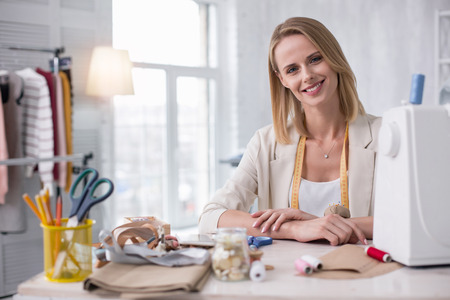 Tailoring practice. Merry female tailor sitting at table while looking at cameraの写真素材