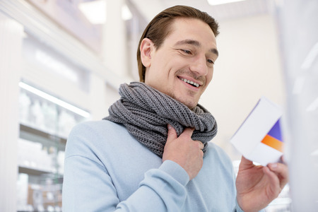 Throat infection. Low angle of vigorous merry man wearing scarf while selecting medication for relieving cold symptomsの写真素材