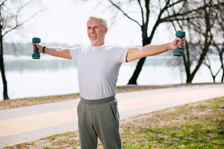Sportive spirit. Vigorous mature man training with dumbbells and standing in park