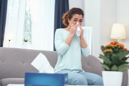 Influenza condition. Adolescent fair haired lady blowing her nose suffering from influenza while staying at home due to her illnessの写真素材