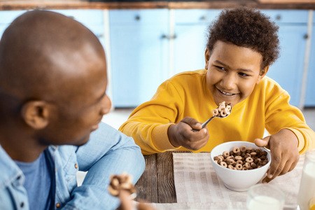 More delicious together. Joyful pre-teen boy sitting at the table next to his father and smiling at his father while eating cereals together with himの写真素材