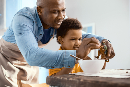 Father-son bond. Charming young father feeding cereals to his little sons toy dinosaur while the boy having breakfast in the kitchenの写真素材