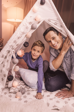 Fun time. Joyful happy nice father and son sitting in the tent and looking at you while playing togetherの写真素材