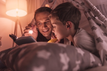 Before sleep. Nice pleasant caring father holding a flash light and smiling while reading a fairy tale to his sonの写真素材