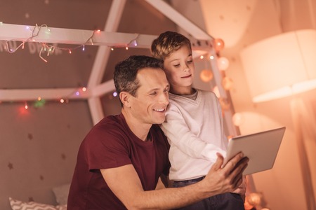 Digital technology. Nice pleasant young boy standing together with his father and pressing his finger to the tablet screen while learning to use modern technologyの写真素材