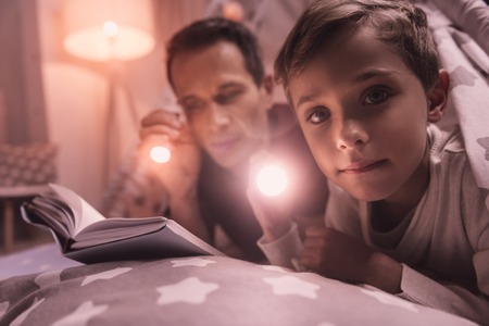 Late at night. Nice pleasant cute boy holding a flash light and looking at you while being with his fatherの写真素材