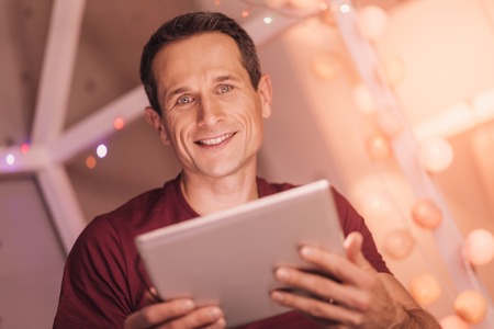 Innovative tablet. Delighted positive handsome man smiling and looking at you while using modern tabletの写真素材