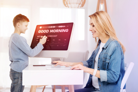 Calm morning. Cheerful creative designer sitting with her tablet and working while her clever smiling son standing next to a modern computerの写真素材