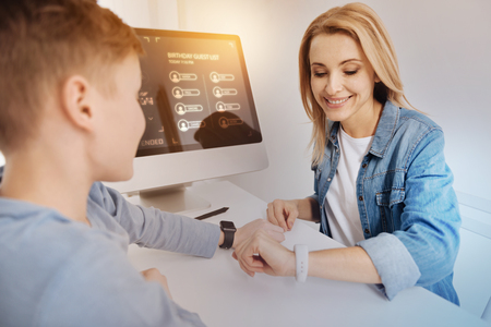 Smart watch. Young emotional mother sitting with her progressive son and smiling while comparing their beautiful convenient smart watchesの写真素材