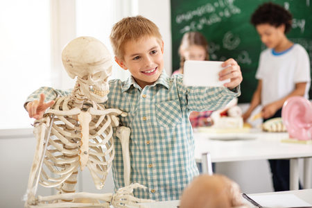 Cool pic. Delighted joyful boy hugging a skeleton while taking a selfie with itの写真素材