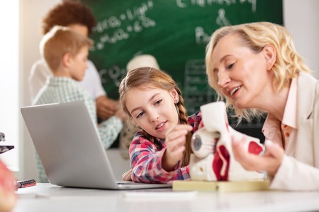 Smart child. Pleasant curious girl sitting near her tutor while trying to touch an eye modelの写真素材