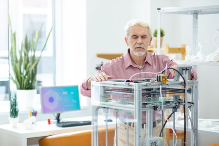 Under control. White-haired elderly man watching a 3D printer work and peeking inside the mechanism