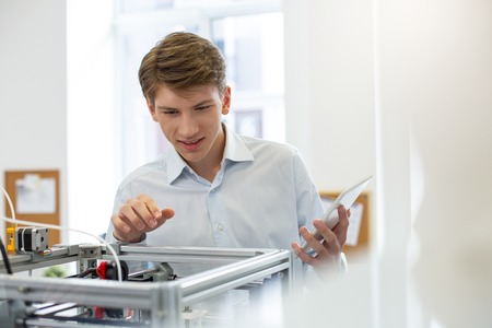 Interesting discovery. Charming young office worker smiling pleasantly while peeking inside 3D printer and discovering something new about its mechanismの写真素材