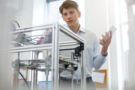 New knowledge. Pleasant young man learning the structure of extruder of a 3D printer while holding a...