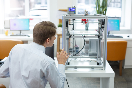 Attentive to details. Charming young office worker looking at the 3D printer and paying attention to...