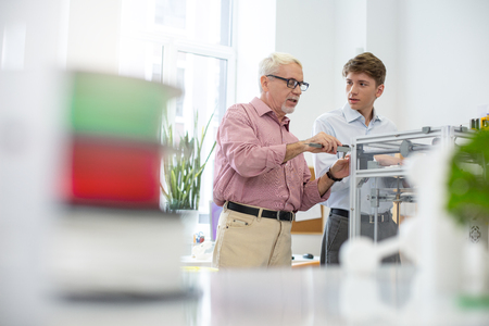 Nice explanation. Charming white-haired man showing his young intern how to use caliper while measuring a model printed with 3D printerの写真素材